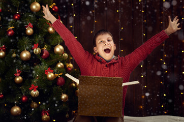 child boy sitting with gift box near christmas decorated fir tree, dark wooden background