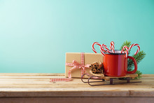 Christmas Background With Cup On Sledges, Candy Cane And Gift Box On Wooden Table
