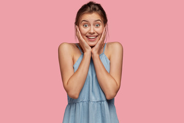 Wall Mural - Joyful young European woman delighted with great result of work, stands happy against pink background, wears denim summer dress, models indoor, listens intrigued story. People and happiness concept