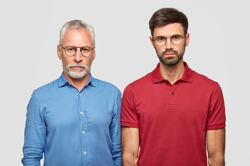 Wall Mural - Father and son have serious expressions, stand closely against white background, have good relationships. Mature handsome man in formal shirt and spectacles stands near his young grandson indoor