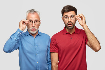 Wall Mural - Waist up shot of two young men of different generation have uexpected gaze at camera, wears round transparent glasses, cant believe in shocking rumors, pose together against white background
