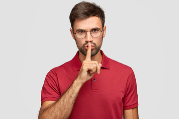 Wall Mural - Shut up, please. Serious unshaven young man wears round transparent glasses, dressed in red casual t shirt, says shh while shows hush gesture, keeps something in secret isolated over white background.