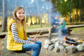 Wall Mural - cute preteen girl roasting hotdog on a stick at bonfire. child having fun at camp fire. camping with