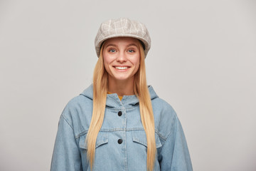 Wall Mural - Close up of a lovely blonde young girl with long hair down, looking happy smiling, wearing oversize denim jacket and beige checked hat cap,isolated over grey background