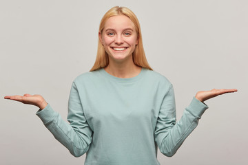 Wall Mural - Close up of a lovely blonde young girl with long hair down, looking happy glad smiling, holding the copyspace on two palms, wearing blue casual sweatshirt,isolated over grey background