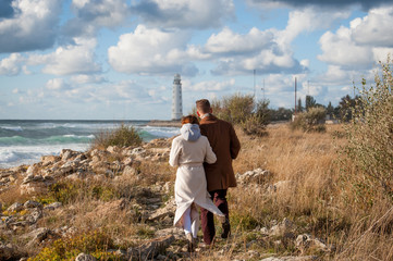 young pair male and woman in coat walking along stormy sea coast towards lighthouse