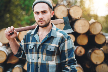 Wall Mural - Strong bearded lumberman holds ax on his shoulder on warehouse of logs. Blurred background