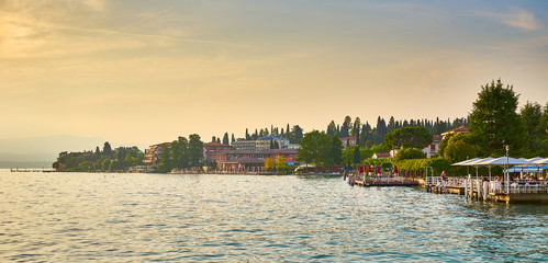 Wall Mural - City of Sirmione at Lake Garda at sunset / People sitting in a restaurant next to harbor with boats 