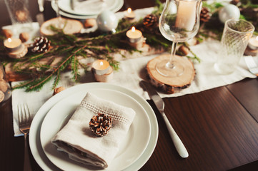 Festive Christmas and New Year table setting in scandinavian style with rustic handmade details in natural and white tones. Dining place decorated with pine cones, branches and candles