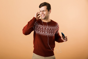 Canvas Print - Happy young man isolated over beige background using phone listening music by earphones singing.