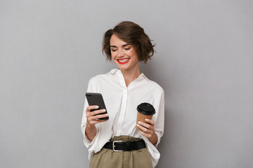 Poster - Photo of european woman 20s holding takeaway coffee and using cell phone, isolated over gray background