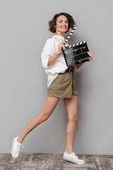 Poster - Photo of brunette woman 20s smiling and holding black clapperboard, isolated over gray background