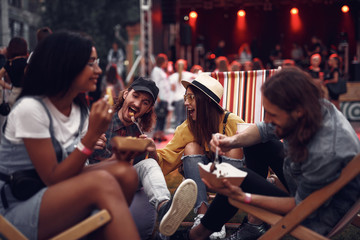 Wall Mural - Portrait of cheerful hipster girl in hat feeding boyfriend with fork while sitting on folding chair. Blurred bearded man and girl in glasses eating and smiling