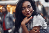 Fototapeta  - Portrait of hipster young lady in glasses holding mobile phone and looking at camera with smile. Street and people on blurred background with bokeh effect
