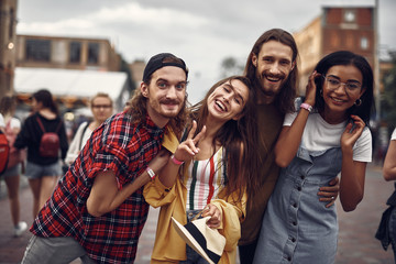 Wall Mural - Portrait of cheerful hipster friends hugging and looking at camera with smiles. Lovely girl holding hat and showing victory gesture