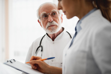 Wall Mural - Portrait of bearded surgeon in glasses staring at young lady in white lab coat while she writing on clipboard. Focus on elderly man