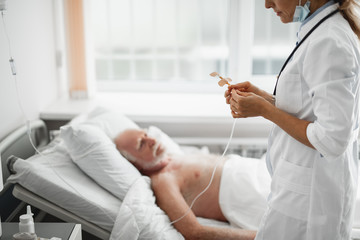 Wall Mural - Cropped side view portrait of young lady in white lab coat checking IV infusion. Old man lying in hospital bed on blurred background