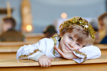 Wall Mural - Beautiful little kid boy playing an angel in Christmas story in a church. Happy adorable blond child with lights and xmas tree on background. Celebration of Xmas eve, big christian, catholic holiday.