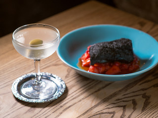 baked pork tenderloin marinated in cuttlefish ink with red tomatos and basil on blue plate and dry vermouth alcohol cocktail with olive on the table in the bar