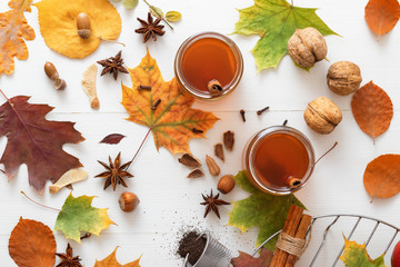 White wooden table with 2 glasses of tea with fall, autumn decoration and white background. Colorful leaves, lemon, cinnamon, walnuts, apples, tea, walnut shells, hazelnuts, tea strainer, acorns.