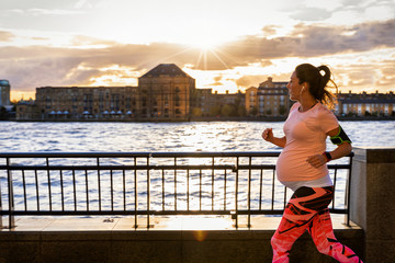 Aktive, schwangere Frau joggt in der Stadt am Flußufer bei Sonnenuntergang