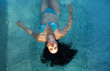 schöne reife Frau im besten Alter mit dunklen Locken im blauen Bikini in der Sonne Sonnenlicht schwebt schwerelos elegant glücklich schwimmend in türkis blauem Wasser im Pool