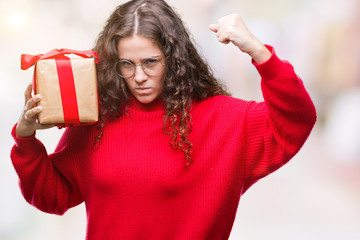 Sticker - Young brunette girl holding a gift over isolated background annoyed and frustrated shouting with anger, crazy and yelling with raised hand, anger concept