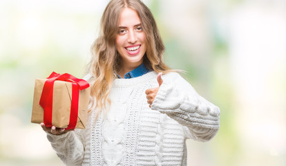 Wall Mural - Beautiful young blonde woman holding gift over isolated background happy with big smile doing ok sign, thumb up with fingers, excellent sign