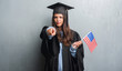 © Krakenimages.com - Young brunette woman over grunge grey wall wearing graduate uniform holding flag of America pointing with finger to the camera and to you, hand sign, positive and confident gesture from the front