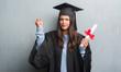 © Krakenimages.com - Young brunette woman over grunge grey wall wearing graduate uniform holding degree annoyed and frustrated shouting with anger, crazy and yelling with raised hand, anger concept