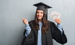 © Krakenimages.com - Young brunette woman over grunge grey wall wearing graduate uniform holding dollars very happy pointing with hand and finger to the side