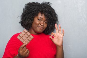 Poster - Young african american woman over grey grunge wall eating a chocolate bar doing ok sign with fingers, excellent symbol