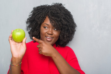 Sticker - Young african american woman over grey grunge wall eating green apple very happy pointing with hand and finger