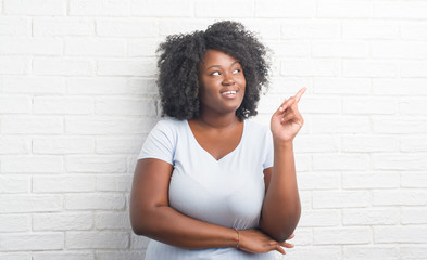 Poster - Young african american plus size woman over white brick wall with a big smile on face, pointing with hand and finger to the side looking at the camera.