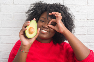 Sticker - Young african american woman over white brick wall eating avocado with happy face smiling doing ok sign with hand on eye looking through fingers