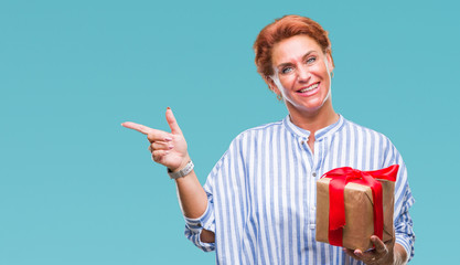 Wall Mural - Senior caucasian woman holding gift over isolated background very happy pointing with hand and finger to the side