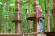 © Tropical studio - Brave little child in safety harness climb on tree tops route to zip line, pass hanging bridge obstacle in adventure rope park. Outdoor activity, family lifestyle in summer camp on vacation with kid