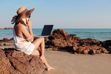 Wall Mural - beautiful and cute businesswoman using laptop computer and cell phone with sun glasses and hat on the beach during holiday vacation