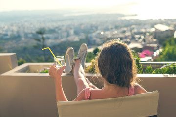 Girl with a long hair in the headphones on the chair on the balcony listening to music on the sunset town background