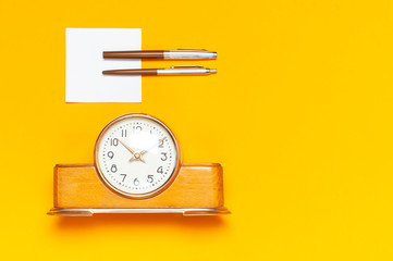 Wooden retro alarm clock, two ballpoint pens and clean white sheets on bright yellow minimalistic background top view Flat Lay with copy space. The concept of time, education.