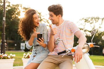 Canvas Print - Portrait of joyful couple man and woman, wearing earphones sitting on scooter together on city street and using smartphone