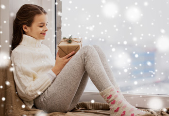 Wall Mural - childhood, winter holidays and people concept - happy beautiful girl with christmas gift sitting on sill at home window