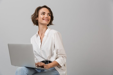 Young business woman posing isolated over grey wall background sitting on stool using laptop computer.