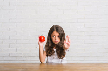 Poster - Young hispanic kid sitting on the table eating fresh tomato with open hand doing stop sign with serious and confident expression, defense gesture