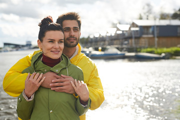 Happy dreamy young couple in coats standing together at seaport and looking at view while contemplating seascape together, smiling man hugging girlfriend