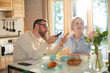 © pressmaster - Young bearded Caucasian man pointing at TV while sitting at kitchen table with his wife, having breakfast and watching morning news attentively