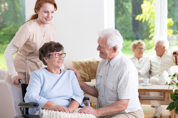 Wall Mural - Smiling elderly man visiting senior woman in the wheelchair while nurse supporting her