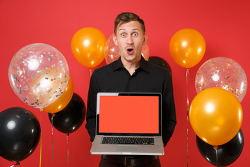 Excited young man in black classic shirt holding laptop pc computer with blank black empty screen on red background air balloons. Valentine's Day, Happy New Year birthday mockup holiday party concept.