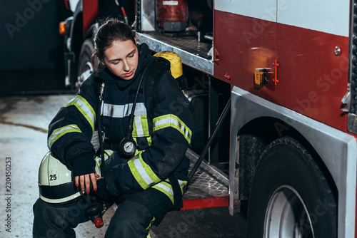 tired female firefighter in uniform with helmet sitting on truck at fire station Poster Mural XXL