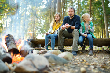 Wall Mural - cute little sisters and their father roasting marshmallows on sticks at bonfire. children having fun
